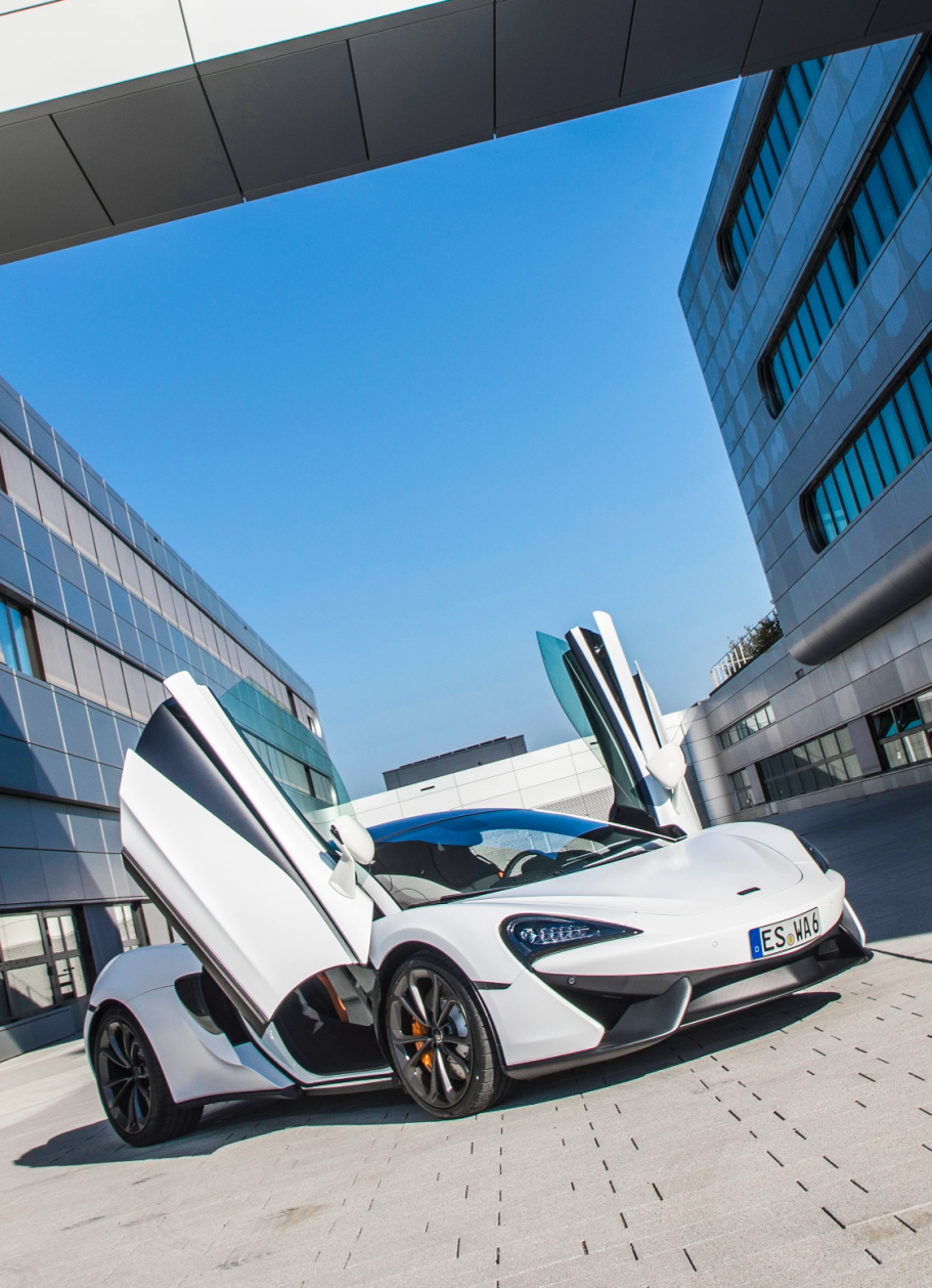 Weißer McLaren 570S mit geöffneten Flügeltüren vor moderner Architektur in dynamischer Perspektive.
