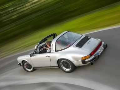 Silberner Porsche 911 Targa in dynamischer Fahrt auf einer kurvenreichen Landstraße.