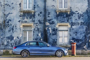 Blaue Mercedes-Benz E‑Klasse Limousine mit AMG-Line-Felgen vor einer blau gestrichenen Fassade.