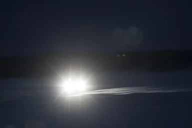 Heller Fahrzeugscheinwerfer in der Nacht mit feinen Lichtspuren auf verschneiter Fahrbahn.