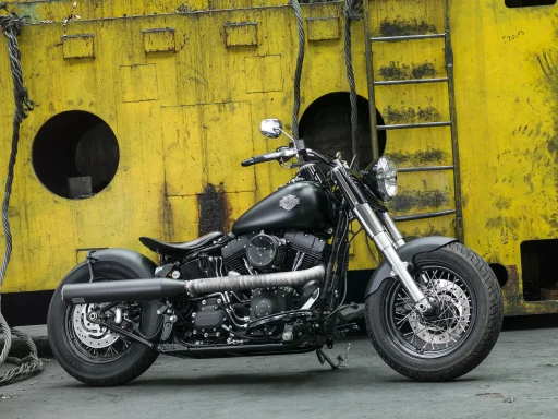 Mattschwarze Harley-Davidson Softail Slim FLS Bobber von 2013 vor einer gelben Industrieanlage mit Leiter und Stahlkonstruktion.