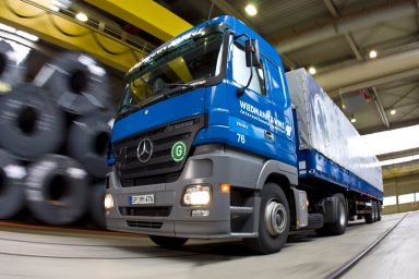 Blauer Mercedes-Benz Actros mit Auflieger fährt durch eine Industriehalle, in der im Hintergrund große Stahlrollen gelagert sind.
