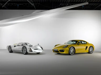 Weißer Porsche 906 Carrera 6 Rennwagen und gelber Porsche Cayman in einem Fotostudio vor weißem Hintergrund.