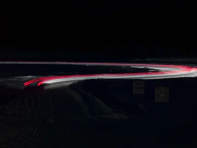 Langzeitbelichtung mit roten und weißen Lichtspuren eines Fahrzeugs auf einer verschneiten Teststrecke bei Nacht.