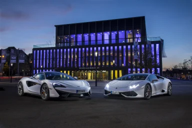 Ein McLaren 570S und ein Lamborghini Huracán, stehen vor einem modernen Gebäude mit blau beleuchteter Glasfassade in der Abenddämmerung.