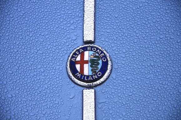 Alfa-Romeo-Emblem auf blauer Motorhaube mit weißen Zierstreifen und Regentropfen.