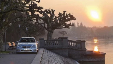 Silberne Mercedes-Benz E‑Klasse W212 an einer Seepromenade bei Sonnenuntergang.