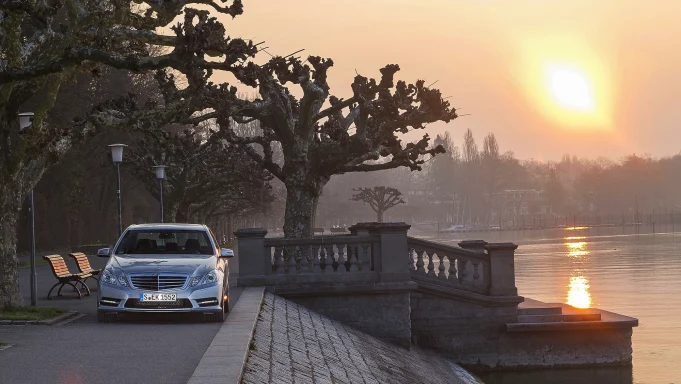 Silberne Mercedes-Benz E‑Klasse W212 an einer Seepromenade bei Sonnenuntergang.