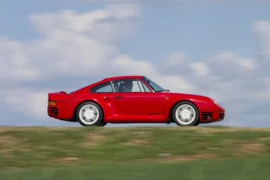 Roter Porsche 959 in Seitenansicht während der Fahrt auf einer Landstraße vor blauem Himmel mit leichter Bewegungsunschärfe im Vordergrund.