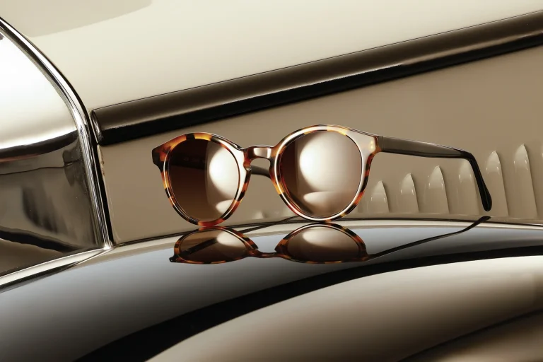 Runde Designerbrille mit blauem Rahmen und gemusterten Bügeln auf der Motorhaube eines Oldtimers neben einem Scheinwerfer.