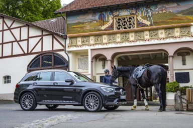 Schwarzer Mercedes-Benz GLC AMG Line vor einem historischen Hofgebäude mit bemalter Fassade. Daneben steht ein gesatteltes schwarzes Pferd.