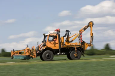 Mercedes-Benz Unimog mit angebautem Mähgerät fährt im Einsatz zur Straßenunterhaltung über eine Landstraße.