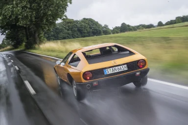 Orangefarbener Mercedes-Benz C111 von hinten auf einer nassen Landstraße in Fahrt, mit sichtbarer Gischt und grüner Landschaft im Hintergrun
