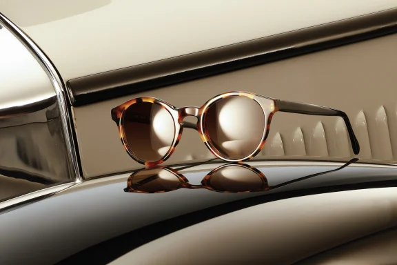 Runde Designerbrille mit blauem Rahmen und gemusterten Bügeln auf der Motorhaube eines Oldtimers neben einem Scheinwerfer.