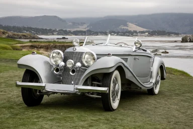 Klassischer silberner Roadster vor einer Küstenlandschaft.