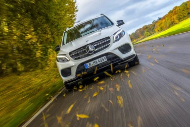 Weißer Mercedes-Benz GLE in Frontansicht während der Fahrt auf einer Landstraße, umgeben von herbstlicher Landschaft und aufgewirbelten Blät