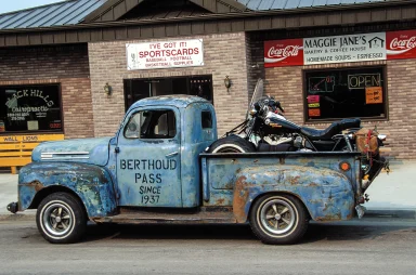Patinierter blauer Ford Pickup mit einer Harley-Davidson auf der Ladefläche vor einem Geschäft in den USA.