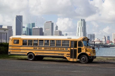 elber amerikanischer Schoolbus steht vor der Skyline von Miami am Ufer einer Wasserfläche.
