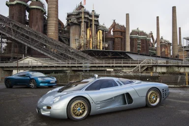 Silbernes Sportauto vor einer industriellen Kulisse mit großen Anlagen im Hintergrund.