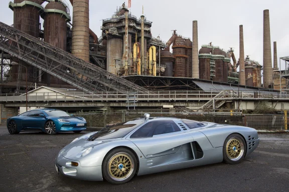 Silbernes Sportauto vor einer industriellen Kulisse mit großen Anlagen im Hintergrund.