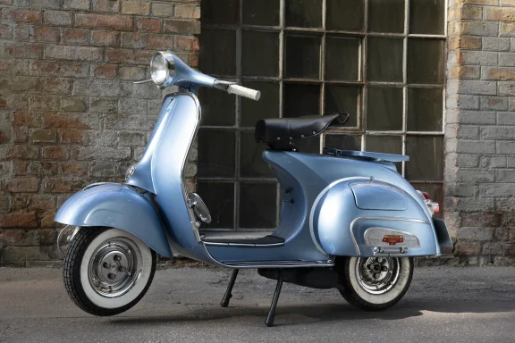 Hellblaue Vespa VBB2T vor einer historischen Backsteinfassade mit Industriefenstern.
