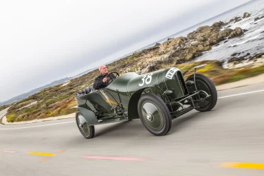 Historischer Prinz-Heinrich-Wagen bei einer dynamischen Fahrt entlang einer Küstenstraße.