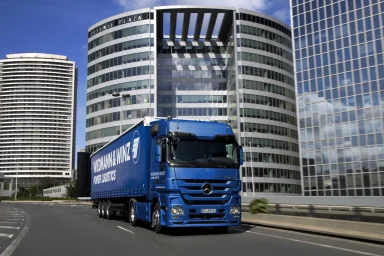 Blauer Mercedes-Benz Actros Sattelzug fährt vor modernen Bürogebäuden in einer Großstadt.