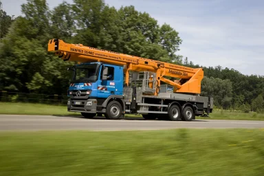Mercedes-Benz Actros mit montierter Arbeitsbühne fährt auf einer Landstraße vor grüner Landschaft.