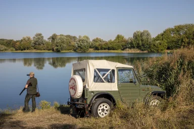 Klassischer Suzuki LJ80 Geländewagen am Seeufer neben einer Person mit Fernglas vor ruhigem Wasser und bewaldeter Landschaft.
