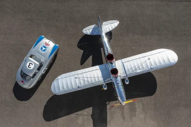 Ein Flugzeug Oldtimer neben einem Rennwagen auf einer asphaltierten Fläche.