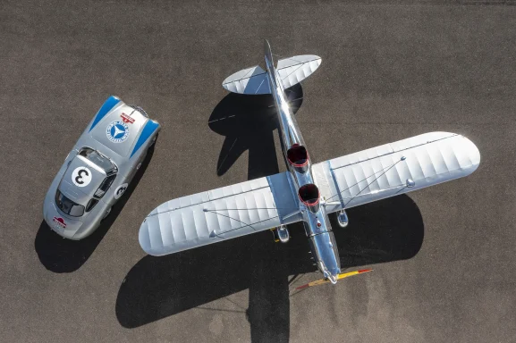 Ein Flugzeug mit breiten Flügeln neben einem Rennwagen auf einer asphaltierten Fläche.