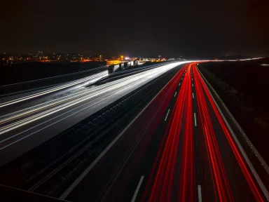 Langzeitbelichtung einer Autobahn bei Nacht mit weißen und roten Lichtspuren vorbeifahrender Fahrzeuge.