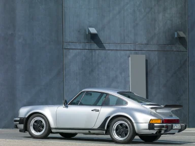 Silberner Sportwagen Porsche 911 Turbo mit markantem Heck und stilvollen Felgen vor grauer Wand.