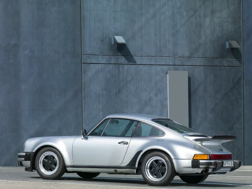 Silberner Sportwagen Porsche 911 Turbo mit markantem Heck und stilvollen Felgen vor grauer Wand.