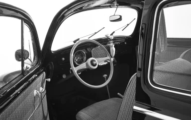 Innenraum eines klassischen Volkswagen Käfers mit weißem Lenkrad, geöffneten Türen und schlichtem Armaturenbrett in Schwarz-Weiß-Darstellung