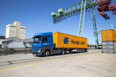 Blauer Mercedes-Benz Actros 1845 transportiert einen Container auf einem Hafenterminal unter einem Portalkran.