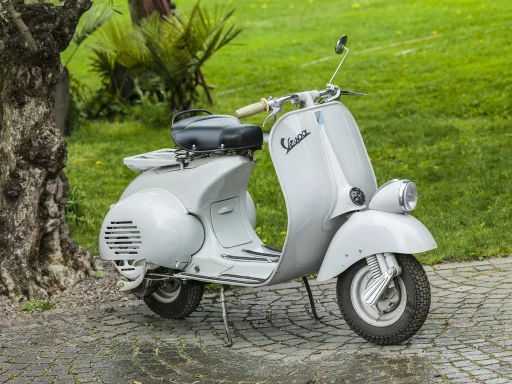 Weiße Vespa V98 Baujahr 1946 auf historischem Kopfsteinpflaster vor grüner Parkkulisse.
