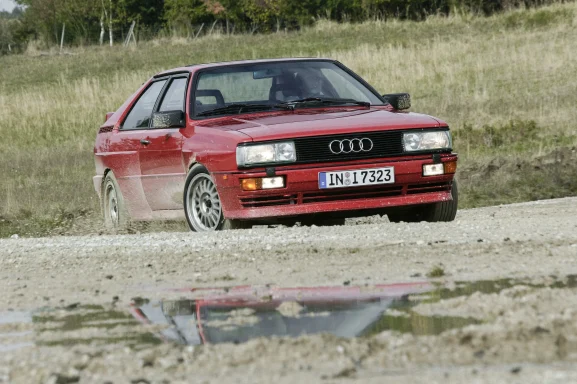 Roter Audi Urquattro in schneller Fahrt auf Schotter mit aufgewirbeltem Kies.
