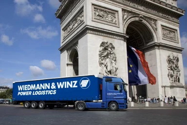 Blauer Lkw der Spedition Wiedmann & Winz fährt vor dem Arc de Triomphe in Paris.
