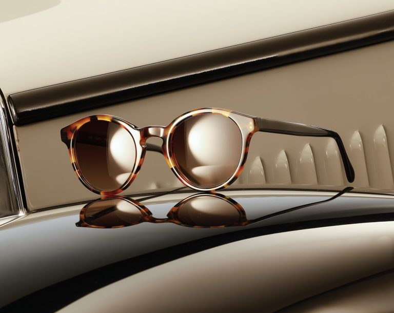 Runde Designerbrille mit blauem Rahmen und gemusterten Bügeln auf der Motorhaube eines Oldtimers neben einem Scheinwerfer.
