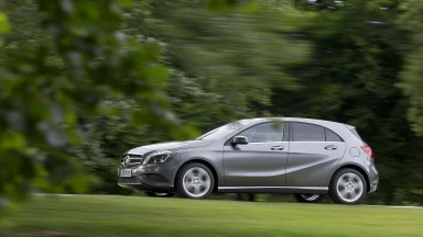 Graue Mercedes-Benz A-Klasse der Baureihe W176 in Seitenansicht während der Fahrt auf einer Straße vor grünem, unscharfem Hintergrund.