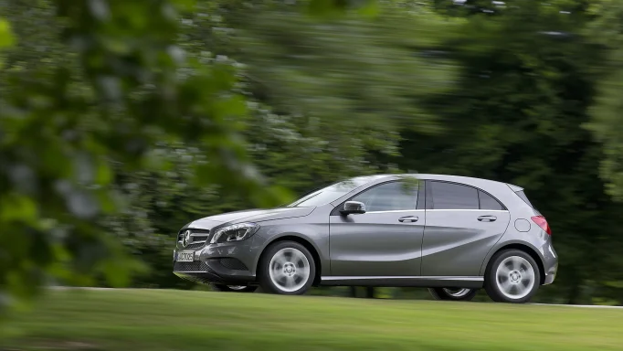 Graue Mercedes-Benz A-Klasse der Baureihe W176 in Seitenansicht während der Fahrt auf einer Straße vor grünem, unscharfem Hintergrund.