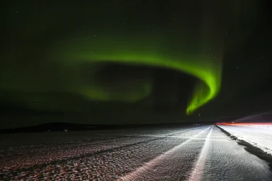 Grünes Polarlicht spannt sich über eine verschneite Straße in Arjeplog, Nordschweden.