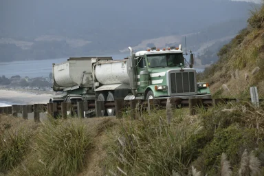 Grüner Peterbilt Kippsattelzug auf einer Küstenstraße in hügeliger Landschaft.