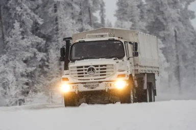 Weißer Mercedes-Benz Xedros Lkw fährt mit eingeschaltetem Licht auf einer verschneiten Straße durch eine winterliche Waldlandschaft.