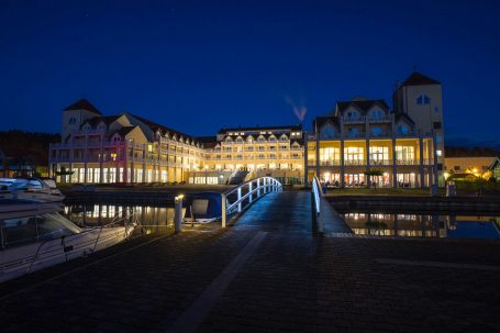Maritim Hafenhotel Rheinsberg bei Nacht mit beleuchteter Fassade und Brücke über dem Wasser.