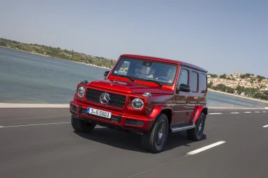 Roter Mercedes-Benz G 500 in Frontansicht während der Fahrt auf einer Küstenstraße mit Blick auf das Meer und eine Ortschaft im Hintergrund.
