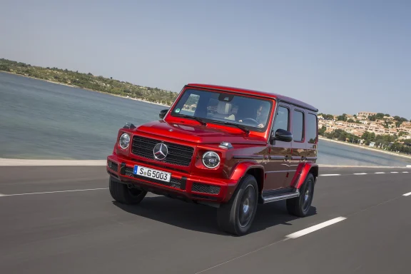 Roter Mercedes-Benz G 500 in Frontansicht während der Fahrt auf einer Küstenstraße mit Blick auf das Meer und eine Ortschaft im Hintergrund.
