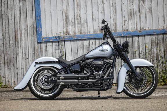 Harley-Davidson Softail im Chicano Style mit silbergrauer Lackierung und großen Speichenrädern vor einer historischen Holzwand.