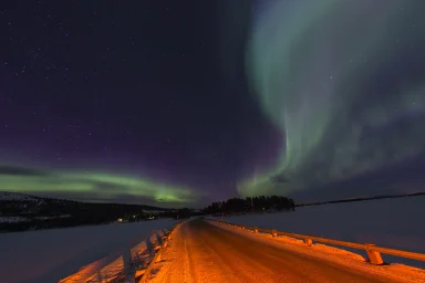 Grüne Polarlichter über einer verschneiten Straße und zugefrorenem See bei Arjeplog in Nordschweden.